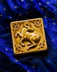 The center of this 4 inch by 4 inch Aries tile features an alert ram with its legs positioned as if it is prancing. The ram's head is turned backwards over its shoulder. The border around the square tile encircles the beast and each of the four corners contains the Aries sign. The tile rests on a deep blue velvet fabric with gold stars speckled across its surface.