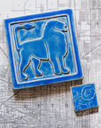 The right-facing Lion tile in the Sky glaze sits next to a three inch by three inch tile featuring a line-drawn image of a cartoonish lion on a black and white map of Detroit. 