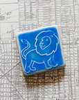 The three inch by three inch Lion tile features a hand-drawn cartoonish lion with a scraggly mane and swishing tail. The tile rests on a black and white map of Detroit.