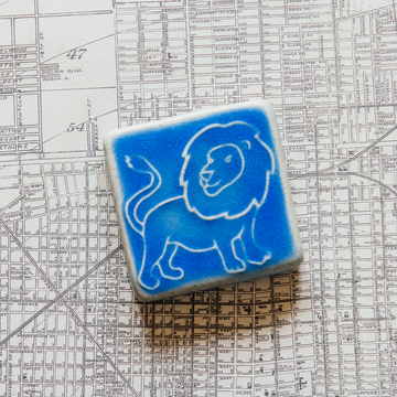 The three inch by three inch Lion tile features a hand-drawn cartoonish lion with a scraggly mane and swishing tail. The tile rests on a black and white map of Detroit.