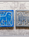 Two 8 inch by 8 inch tiles sit on a black and white map of Detroit. Each tile features a simplified silhouette of a lion's body, each of their tails are alert and pointed forward to their head. The tiles are mirrored, with the bright blue tile on the left facing the gray-blue tile on the right. 