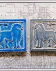 Two 8 inch by 8 inch tiles sit on a black and white map of Detroit. Each tile features a simplified silhouette of a lion's body, each of their tails are alert and pointed forward to their head. The tiles are mirrored, with the bright blue tile on the left facing the gray-blue tile on the right.