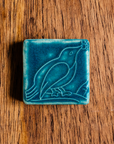The ceramic Bird with Berries Tile depicts a bird sitting on a branch with berries in its beak. This tile is glazed in a matte Peacock blue color.