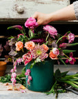 This Pewabic Blue crock sits outside on a wooden work table covered in petals and garden snips. A hand reaches into frame to place a flower, just plucked from the garden into the mostly finished bouquet.