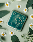 The Cactus Flower Tile features a large, four petalled flower on a thicker cactus stem. Two small petals seem to fly in the wind next to it. This tile features the medium blue Glacier Gloss glaze.