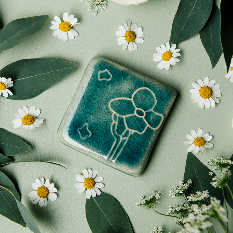 The Cactus Flower Tile features a large, four petalled flower on a thicker cactus stem. Two small petals seem to fly in the wind next to it. This tile features the medium blue Glacier Gloss glaze.