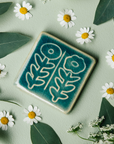 The Two Flowers Tile features two flowers on long, straight, leafy stems. One flower has scalloped petals while the other is circular. This tile features the medium blue Glacier Gloss glaze.