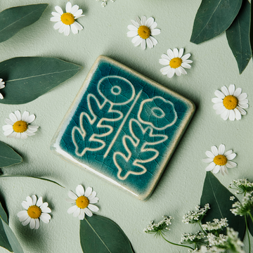 The Two Flowers Tile features two flowers on long, straight, leafy stems. One flower has scalloped petals while the other is circular. This tile features the medium blue Glacier Gloss glaze.
