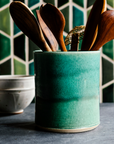 This Pewabic green Crock is holding wooden cooking utensils on a kitchen counter. The backsplash features multicolored green Pewabic tiles in a hexagonal design.