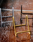 All three matte gold, silver, and bronze tile stands photographed together on a tiled-window ledge.