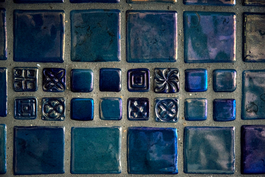 A detail of a tile installation showcases the blues and purples of a metallic iridescent glaze. Some of the smaller square tiles are embossed with geometric designs.