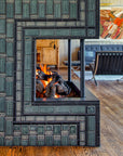 Side view of the lit fireplace. Two mid-century modern chairs and a cozy living room area are visible in the frame. The natural-finished wood vaulted ceiling is a beautiful contrast to the strong geometric design of the fireplace installation.