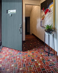 A door opens up to a tiled foyer comprised of warm, red and earth toned tiles with bright, luminous "blush" iridescent tiles that look golden with the sunshine creeping in. There is a large mirror-like charcoal glazed Pewabic vase with a plant on a stand in-frame against thew wall. The photo is taken at an angle to the right of the open door.