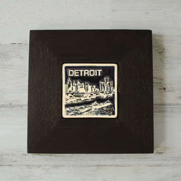The Detroit Skyline tile features the current downtown skyline with the Detroit River in the foreground. A freighter floats by causing ripples on the otherwise flat surface of the water. The word "Detroit" is written above in the sky. The Skyline buildings, freighter and word are glazed in a glossy white while the background is glazed in a deep black. The wooden frame is matte black.