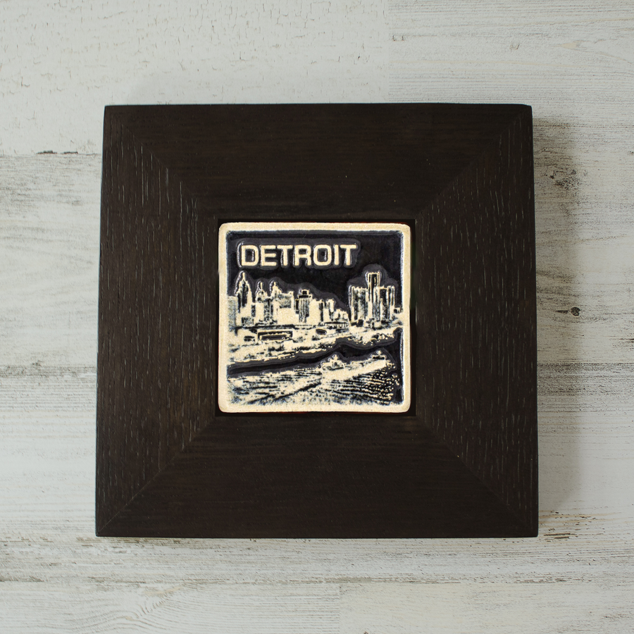 The Detroit Skyline tile features the current downtown skyline with the Detroit River in the foreground. A freighter floats by causing ripples on the otherwise flat surface of the water. The word "Detroit" is written above in the sky. The Skyline buildings, freighter and word are glazed in a glossy white while the background is glazed in a deep black. The wooden frame is matte black.