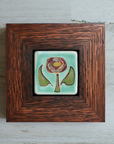 The Hand-Painted Rose Tile features a line drawing of a pinkish dusty rose in the center with a green stem and leaves on a pale blue background. The frame is a deep reddish brown oak wood frame.
