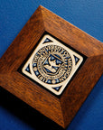 A 4x4 Midnight Scraped University of Michigan Seal tile in a rich wood frame rests on a royal blue surface