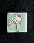 The Hand-Painted Crane Tile features a line drawing of a white crane with its whole body turned to the right, one leg on the ground and the other folded up, pointing to the right. The background is a pale greenish-blue.