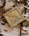 The Dragonfly Tile features a detailed dragonfly with wings outstretched. It is sitting horizontally on the square tile with a thick border around the edge. The pinkish gold metallic Blush Iridescent glaze features lots many variations. Depending on the lighting in the room, these pieces will pick up different hues.