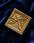 The Dragonfly Tile features a detailed dragonfly with wings outstretched. It is sitting horizontally on the square tile with a thick border around the edge. The pinkish gold metallic Blush Iridescent glaze features lots many variations. Depending on the lighting in the room, these pieces will pick up different hues.