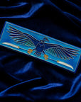 The short and wide Nekhbet tile features a line drawing of a vulture with wings outstretched, head turned to the right as she holds two stylized feathers- one in each talon. This tile features bright shades of blue. This tile rests on a bed of rich, blue velvet.