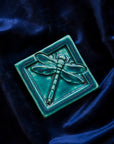The Dragonfly Tile features a detailed dragonfly with wings outstretched. It is sitting horizontally on the square tile with a thick border around the edge. This Dragonfly Tile features the blueish-teal Pewabic Blue glaze.