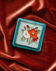 A Hand-Painted Bouquet Tile in our "Poppy Palette" rests on a bed of rich, organge-pink velvet.