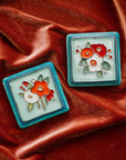 A Hand-Painted Three Flowers Tile and Hand-Painted Bouquet Tile both in our "Poppy Palette" rests on a bed of rich, organge-pink velvet.