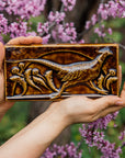 The long, horizontal Pheasant Tile features a male ring-necked pheasant running across the tiles. Puffy late-summer dandelions surround its outstretched feet. A large, domed sun is setting behind it. Featured in in a glossy brown Molasses glaze.