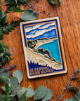 The Sleeping Bear Dunes Tile features the serene Lake Michigan and the sandy beaches and dunes covered on top by bushes and greenery. The words "Sleeping Bear Dunes" line the bottom of the design. The hand painted tile boasts blues and greens and pale yellow sand.