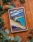 The Sleeping Bear Dunes Tile features the serene Lake Michigan and the sandy beaches and dunes covered on top by bushes and greenery. The words "Sleeping Bear Dunes" line the bottom of the design. The hand painted tile boasts blues and greens and pale yellow sand.