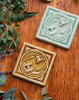 A pair of Daffodil Tiles rest on a beautiful wood finish surrounded by summer greenery. They are glazed in our glossy Celadon and Honey glaze.