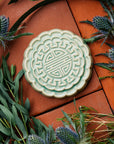A Mooncake Paperweight in our glossy Celadon glaze, surrounded by thistle and wax flowers.