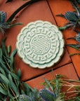 A Mooncake Paperweight in our glossy Celadon glaze, surrounded by thistle and wax flowers.
