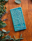 This Pewabic Blue Detroit Map Tile is sitting on a wooden board, surrounded by flowers and eucalyptus.