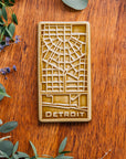 This Honey Glazed Detroit Map Tile is sitting on a wooden board, surrounded by flowers and eucalyptus.