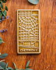 This Honey Glazed Detroit Map Tile is sitting on a wooden board, surrounded by flowers and eucalyptus.