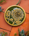 A two-tone glossy, round Sunflower Trivet with golden border and outline set against an emerald green background.