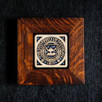 A 4x4 Midnight Scraped University of Michigan Seal tile in a rich wood frame rests on a black surface