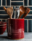 This crock holds wooden cooking utensils on a kitchen counter. The backsplash is decked with long rectangular Pewabic tiles in a deep, grayish green color.