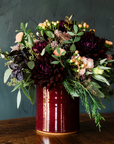 The crock is a cylindrical vessel with straight sides that has a wide opening and is as wide as it is tall. This crock is filled with a large bouquet of burgandy and white flowers.