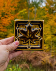 A person's hand is reaching into the bottom-left of the frame holding a Maple Leaf tile in a deep, glossy Molasses glaze. There are trees in the background with vibrant, autumn leaves. 