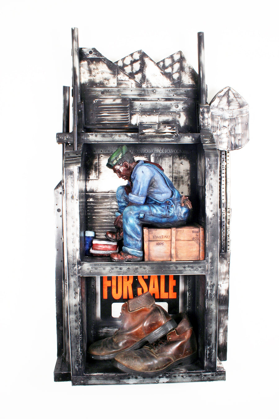 The top shelf is empty with harshly shaped metal that gives the impression of mountains at its top. The middle shelf holds a ceramic figure of a black man in denim work overalls and a green hat sitting on a wooden box as he ties his work boots. His shoe is resting on his lunch cooler that sits next to a thermos. The bottom shelf features a For Sale sign erected behind a pair of worn leather work boots.