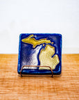 The Michigan Outline Tile features the outline of the state of Michigan with both peninsulas and its larger northern islands. This hand painted tile has a background of deep matte blue while the peninsulas are a maize gold color.