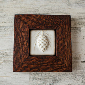 The ceramic Pinecone Tile has a high relief design with one large pinecone in the center and long spindly pine needles framing it. This tile is in the satin finished white Alabaster glaze which beautifully offsets the deep reddish brown hue of the oak frame.