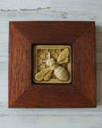 The Honeybee Tile features a large honeybee with stripes and detailed segmented wings. The bee sits on a honeycomb patterned background. The tile is in our deep golden Honey Gloss glaze which is beautifully offset by the deep reddish brown oak wood frame.