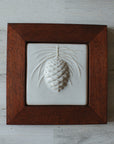 The ceramic Pinecone Tile has a high relief design with one large pinecone in the center and long spindly pine needles framing it. This tile is in the satin finished white Alabaster glaze which beautifully offsets the deep reddish brown hue of the oak frame.