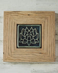 The Lotus tile features a line drawing of a blooming lotus flower, its pointed petals outstretching. It has a simple border around the design. A pale reclaimed wood frame holds the tile in the glossy deep blue Ocean glaze.