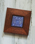 The Scarab Tile features a line drawing of a scarab beetle with wings outstretched and two small circles floating between its front arms. The design has classic Egyptian markings. This tile is in the glossy bright blue Lake Superior glaze which beautifully offsets the deep reddish brown oak wood frame.