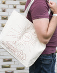 This cream canvas tote bag features a line drawing of the Tudor-style Pewabic building with trees around it, in the sky is a huge image of the sun. It has softly closed eyes and a nose with long rays emanating from it.